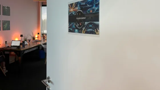 Geöffnete Tür mit einem Schild ‚Kreativlabor‘, das ein Bild eines Autoinnenraums zeigt. Dahinter ein schwach beleuchtetes Büro mit Schreibtisch, Laptop und Pflanzen.