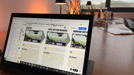 Laptop auf einem Tisch mit geöffneter Miro-Board-Präsentation, die vier Bilder eines Autoinnenraums zeigt. Beschriftungen: ‚Nähernd‘, ‚Kurz vor dem Bremsen‘, ‚Während der Bremsung‘. Im Hintergrund ein Büro mit Pflanzen, Lampen und Getränken.