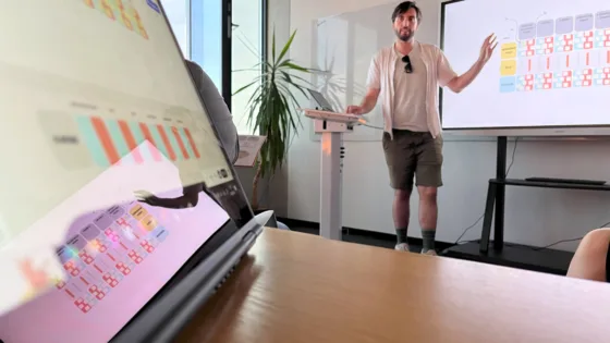 Ein Mann präsentiert vor einem Bildschirm mit farbigen Diagrammen und Tabellen. Im Vordergrund ein Laptop mit derselben Präsentation. Im Hintergrund eine Pflanze neben einem Fenster.“
