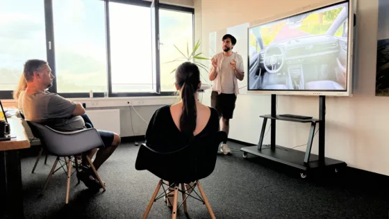 Ein Mann präsentiert vor einem Bildschirm mit dem Bild eines Autoinnenraums. Zwei Personen sitzen und hören zu. Im Hintergrund sind Fenster, eine Pflanze und ein Whiteboard sichtbar.