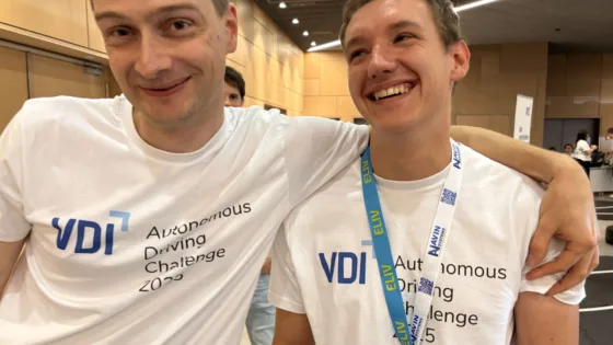Zwei Teilnehmende des Teams Köln, der VDI Autonomous Driving Challenge 2025, powered by MdynamiX. Beide tragen weiße Event-T-Shirts mit der Aufschrift „VDI Autonomous Driving Challenge 2025“ und blaue Lanyards mit dem Schriftzug „ELIV“. Eine Person hat den Arm locker um die Schulter der anderen gelegt. Im Hintergrund sind weitere Personen und Präsentationsflächen sichtbar. Die Szene vermittelt Teamgeist und lockere Atmosphäre während des Wettbewerbs.