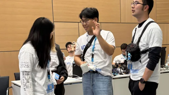Teammitglieder der TU Berlin bei der VDI Autonomous Driving Challenge 2025 powered by MdynamiX bei der ELIV in Bonn stehen zusammen und besprechen sich. Sie tragen weiße T-Shirts mit dem Event-Logo und arbeiten an einem Tisch, auf dem ein schwarzes autonomes Modellfahrzeug platziert ist.