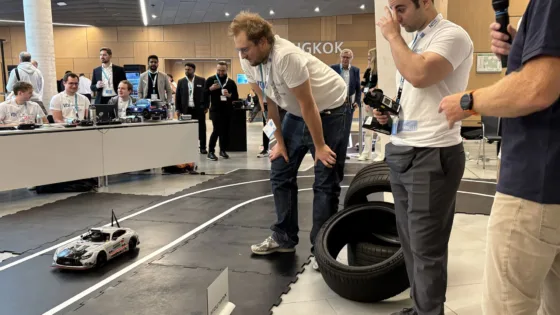 Autonomes Modellfahrzeug der MdynamiX, das MXcarkit, in grauer Rennoptik fährt auf einer markierten Indoor-Teststrecke bei der VDI Autonomous Driving Challenge 2025 powered by MdynamiX auf der ELIV in Bonn. Rechts im Bild liegen mehrere Reifen als Hindernisse. Im Vordergrund stehen die Teilnehmer des Team TU Berlin mit Event-T-Shirts und Kameras, die den Lauf beobachten. Im Hintergrund sitzen weitere Teilnehmer an Tischen mit Laptops, während andere den Wettbewerb verfolgen.