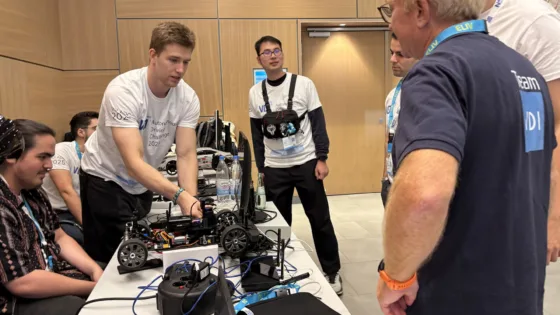 Team der TU Berlin bereitet ein autonomes Modellfahrzeug bei der VDI Autonomous Driving Challenge 2025 powered by MdynamiX auf der ELIV in Bonn vor. Das Fahrzeug steht auf einem Tisch, umgeben von Kabeln, Elektronik und Werkzeugen. Mehrere Personen, darunter die Teammitglieder selbst in weißen Event-T-Shirts und dem Juror Markus Krug, mit VDI-Team-Shirt stehen um den Tisch und arbeiten gemeinsam an der Technik.