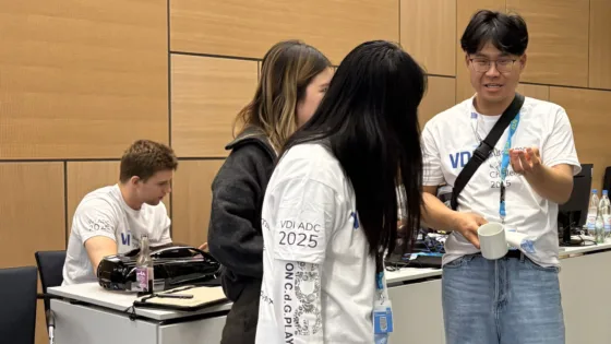 Teammitglieder der TU Berlin bei der VDI Autonomous Driving Challenge 2025 powered by MdynamiX bei der ELIV in Bonn stehen zusammen und besprechen sich. Sie tragen weiße T-Shirts mit dem Event-Logo und arbeiten an einem Tisch, auf dem ein schwarzes autonomes Modellfahrzeug platziert ist.