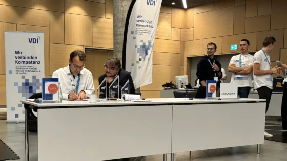 Jurymitglieder und Teilnehmende bei der VDI Autonomous Driving Challenge 2025, powered by MdynamiX. Im Vordergrund steht ein Tisch mit Schildern „JURY“ und „MdynamiX“, mehreren Pokalen und einer kleinen Ampel als Dekoration. Dahinter sind VDI-Banner mit der Aufschrift „Wir verbinden Kompetenz“ sowie ein Reifenstapel sichtbar. Im Hintergrund befinden sich weitere Personen in Event-T-Shirts und eine moderne Halle mit Holzverkleidung und Technik-Equipment. Die Szene vermittelt den organisatorischen Bereich des Wettbewerbs.