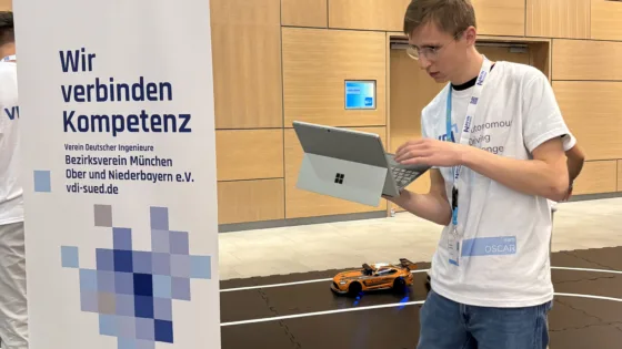 Teilnehmer der VDI Autonomous Driving Challenge 2025, powered by MdynamiX, steht auf einer schwarzen Teststrecke mit weißen Linien in einer modernen Halle. Die Person hält ein silbernes Microsoft Surface-Tablet und trägt ein weißes Event-T-Shirt mit der Aufschrift „VDI Autonomous Driving Challenge 2025“ sowie ein Namensschild „Team OSCAR“. Im Vordergrund links ist ein Banner mit der Aufschrift „Wir verbinden Kompetenz – Verein Deutscher Ingenieure Bezirksverein München Ober- und Niederbayern e.V. vdi-sued.de“ sichtbar. Im Hintergrund fäh