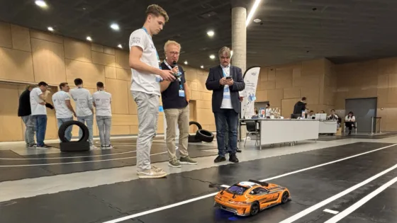 Testszene bei der VDI Autonomous Driving Challenge 2025, powered by MdynamiX, auf der ELIV in Bonn. Im Vordergrund fährt ein orangefarbenes Modellfahrzeug mit Antennen auf einer schwarzen Teststrecke mit weißen Linien. Daneben stehen drei Personen in weißen Event-T-Shirts und einem dunklen Sakko, die offenbar die Fahrzeugbewegung überwachen oder steuern. Im Hintergrund sind weitere Teilnehmende in Gruppen, Reifenstapel als Hindernisse und Präsentationstische sichtbar. Die Umgebung ist eine moderne Halle mit Holzverkleidung und klarer Beleuchtung.