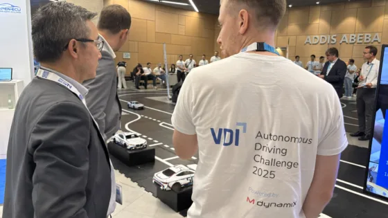 Teilnehmende und Gäste beobachten die das Geschehen der VDI Autonomous Driving Challenge 2025 bei der ELIV in Bonn. Im Vordergrund ist die Rückseite eines weißen T-Shirts mit dem VDI-Logo und der Aufschrift ‚Autonomous Driving Challenge 2025 powered by MdynamiX‘ zu sehen. Auf der Strecke fahren mehrere autonome Modellfahrzeuge, während im Hintergrund weitere Personen den Wettbewerb verfolgen. 