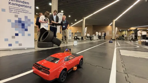 VDI Autonomous Driving Challenge 2025, powered by MdynamiX, auf der ELIV in Bonn. Im Vordergrund fährt ein rotes Modellfahrzeug mit Antennen auf einer schwarzen Teststrecke mit weißen Linien. Die Strecke ist mit Reifenstapeln als Hindernisse ausgestattet. Links steht ein Banner mit der Aufschrift „Wir verbinden Kompetenz“ und der VDI-Website „vdi-sued.de“. Im Hintergrund befinden sich mehrere Personen, darunter die Teilnehmenden der HS München sowie die Jury in weißen Event-T-Shirts, die sich um die Strecke versammelt haben, sowie Präsentationstische und technische Geräte. Die Umgebung ist eine moderne Halle mit Holzverkleidung und klarer Beleuchtung.