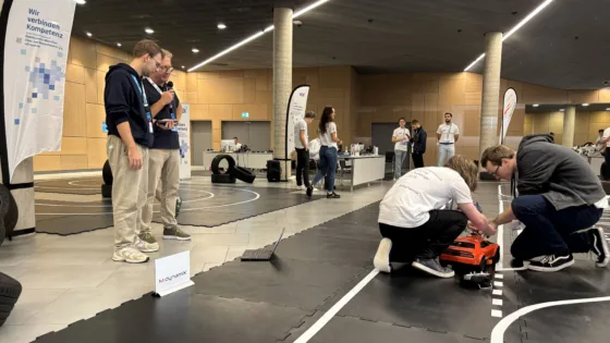 VDI Autonomous Driving Challenge 2025, powered by MdynamiX, auf der ELIV in Bonn. Im Vordergrund arbeiten zwei Personen des Teams der HS München an einem roten Modellfahrzeug auf einer schwarzen Teststrecke mit weißen Linien. Daneben liegt ein geöffneter Laptop. Links im Bild steht ein Banner mit der Aufschrift „Wir verbinden Kompetenz“ und der VDI-Website „vdi-sued.de“. Im Hintergrund sind weitere Teilnehmende in weißen Event-T-Shirts sowie Reifenstapel und Präsentationstische sichtbar. Die Umgebung ist eine moderne Halle mit Holzverkleidung und klarer Beleuchtung.