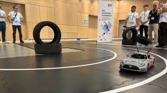 Fahrszene bei der VDI Autonomous Driving Challenge 2025, powered by MdynamiX, auf der ELIV in Bonn. Im Vordergrund fährt ein silbernes Modellfahrzeug, das MXcarkit, mit Antennen auf einer schwarzen Teststrecke mit weißen Markierungen. Auf der Strecke sind mehrere Reifenstapel als Hindernisse platziert. Im Hintergrund stehen Teilnehmende in weißen Event-T-Shirts neben einem VDI-Banner mit der Aufschrift „Wir verbinden Kompetenz“. Die Umgebung ist eine moderne Halle mit Holzverkleidung und technischer Ausstattung.