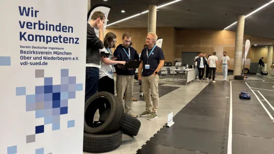 VDI Autonomous Driving Challenge 2025, powered by MdynamiX, auf der ELIV in Bonn. Im Vordergrund fährt ein rotes Modellfahrzeug mit Antennen auf einer schwarzen Teststrecke mit weißen Linien. Die Strecke ist mit Reifenstapeln als Hindernisse ausgestattet. Links steht ein Banner mit der Aufschrift „Wir verbinden Kompetenz“ und der VDI-Website „vdi-sued.de“. Im Hintergrund befinden sich mehrere Personen, darunter die Teilnehmenden der HS München sowie die Jury in weißen Event-T-Shirts, die sich um die Strecke versammelt haben, sowie Präsentationstische und technische Geräte. Die Umgebung ist eine moderne Halle mit Holzverkleidung und klarer Beleuchtung.