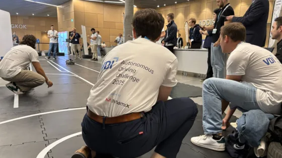 VDI Autonomous Driving Challenge 2025 bei der ELIV in Bonn beobachten ein Beschleunigungsrennen auf der Indoor-Teststrecke. Im Vordergrund knien Teilnehmer in weißen Event-T-Shirts mit der Aufschrift ‚Autonomous Driving Challenge 2025 powered by MdynamiX‘. Sie sind bereit das beschleunigende Fahrzeug am Ende der Strecke zu fangen, sollte es ausbrechen oder nicht rechtzeitig zum Stehen kommen. Auf der Strecke fährt ein autonomes Modellfahrzeug, während im Hintergrund weitere Teams und Zuschauer den Wettbewerb verfolgen. 
