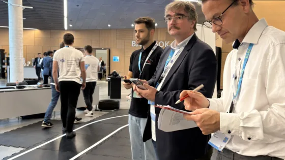 Jury-Mitglieder der VDI Autonomous Driving Challenge 2025 powered by MdynamIX bei der ELIV in Bonn stehen an der Indoor-Teststrecke und machen Notizen sowie Bewertungen. Im Vordergrund sind ein Klemmbrett, ein Stift und ein Smartphone sichtbar. Im Hintergrund laufen Teilnehmer in weißen Event-T-Shirts auf der markierten Strecke, auf der autonome Modellfahrzeuge getestet werden.
