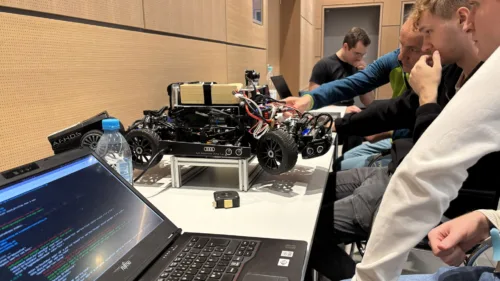 Eine Szene der VDI Autonomous Driving Challenge 2025 bei der ELIV in Bonn. Auf einem Tisch steht ein Modellfahrzeug, das mit zahlreichen Kabeln, Sensoren und elektronischen Komponenten ausgestattet ist und ein Audi-Logo trägt. Daneben liegt ein Laptop mit geöffnetem Code-Editor, auf dem farbiger Programmcode sichtbar ist. Im Hintergrund sind mehrere Personen in eine Diskussion vertieft, während eine Hand am Fahrzeug arbeitet.