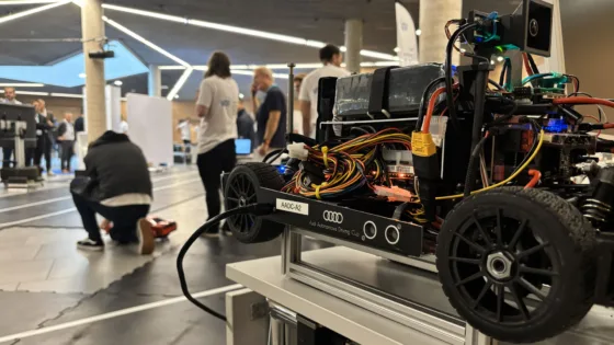 Detailaufnahme eines autonomen Modellfahrzeugs bei der VDI Autonomous Driving Challenge 2025 auf der ELIV in Bonn. Das Fahrzeug steht ohne Karosserie auf einem Podest und zeigt Einblick in das Innenleben, darunter komplexe Verkabelung, Sensoren und Elektronikkomponenten. Auf der Seite sind Logos von Audi und der Challenge sichtbar. Im Hintergrund sind mehrere Personen und eine Indoor-Teststrecke mit markierten Fahrspuren zu erkennen.