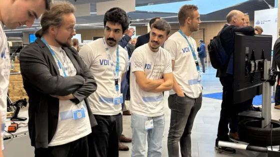 Team Bochum bei der VDI Autonomous Driving Challenge 2025, powered by MdynamiX, stehen in einer modernen Halle vor einem großen Monitor. Alle tragen weiße Event-T-Shirts mit der Aufschrift „VDI Autonomous Driving Challenge 2025“ und blaue Lanyards. Im Vordergrund sind technische Geräte wie ein Modellfahrzeug und Kabel sichtbar. Im Hintergrund befinden sich weitere Personen in Business-Kleidung sowie Präsentationsstände. Die Szene vermittelt konzentrierte Beobachtung und Analyse während des Wettbewerbs.