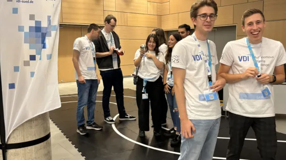 Zu sehen ist das Team der HS München bei der VDI Autonomous Driving Challenge 2025, powered by MdynamiX. Alle tragen weiße Event-T-Shirts mit der Aufschrift „VDI ADC 2025“ und blaue Lanyards. Im Vordergrund ist ein Banner mit der VDI-Website „vdi-sued.de“ sichtbar. Die Gruppe wirkt ausgelassen und freudig.