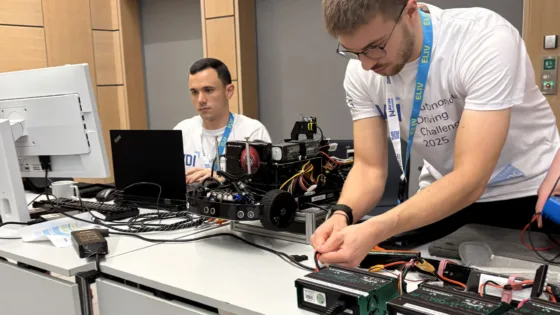 Teilnehmende der VDI Autonomous Driving Challenge 2025, powered by MdynamiX, arbeiten an einem Modellfahrzeug auf einem Tisch. Das Fahrzeug ist mit zahlreichen Kabeln, Sensoren und elektronischen Komponenten ausgestattet. Auf dem Tisch liegen mehrere Akkus, Kabel und Werkzeuge. Im Hintergrund steht ein Monitor und ein Laptop, die für Programmierung oder Tests genutzt werden. Die Szene vermittelt konzentrierte technische Arbeit und Vorbereitung im Rahmen des Wettbewerbs.