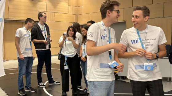 Zu sehen ist das Team der HS München bei der VDI Autonomous Driving Challenge 2025, powered by MdynamiX. Alle tragen weiße Event-T-Shirts mit der Aufschrift „VDI ADC 2025“ und blaue Lanyards. Im Vordergrund ist ein Banner mit der VDI-Website „vdi-sued.de“ sichtbar. Die Gruppe wirkt ausgelassen und freudig.