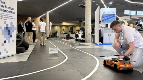 Team Osnabrück bei der VDI Autonomous Driving Challenge 2025, powered by MdynamiX. Im Vordergrund wird ein orangefarbenes Modellfahrzeug auf einer schwarzen Teststrecke mit weißen Linien positioniert. Die Strecke ist mit Schildern wie „PIT OUT“ und Reifenstapeln als Hindernisse ausgestattet. Links steht ein Banner mit der Aufschrift „Wir verbinden Kompetenz“ und der VDI-Website „vdi-sued.de“. Im Hintergrund sind weitere Teilnehmende in weißen Event-T-Shirts sowie Präsentationsstände mit Technik und Monitoren sichtbar. Die Umgebung ist eine moderne Halle mit klarer Beleuchtung und strukturierter Fläche.