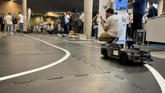 ndoor-Teststrecke bei der VDI Autonomous Driving Challenge 2025 auf der ELIV in Bonn. Im Vordergrund fährt ein schwarzes Modellauto mit Sensoren und Kameras auf einer markierten Fahrbahn. Im Hintergrund stehen mehrere Teams in weißen VDI-T-Shirts sowie weitere Teilnehmer und Beobachter, die die Fahrzeuge und den Wettbewerb verfolgen. Auf der Strecke sind weitere autonome Modellfahrzeuge zu sehen, umgeben von Messgeräten und Laptops.“