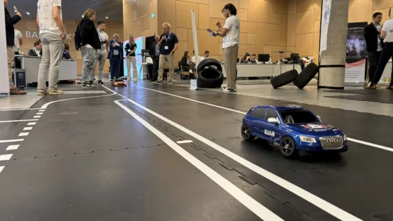 Autonomes Modellfahrzeug in blauer SUV-Optik fährt auf einer markierten Indoor-Teststrecke bei der VDI Autonomous Driving Challenge 2025 auf der ELIV in Bonn. Im Hintergrund stehen mehrere Personen, die den Wettbewerb beobachten oder Fahrzeuge vorbereiten. Auf der Strecke sind Hindernisse wie Reifen platziert, die Teil der dynamischen Disziplin ‚am Hindernis vorbeifahren‘ sind.