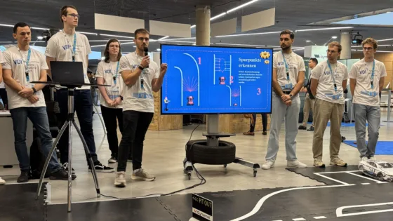 Präsentation der HS München bei der VDI Autonomous Driving Challenge 2025, powered by MdynamiX. Eine Gruppe von Teilnehmenden steht auf einer markierten Teststrecke neben einem großen Bildschirm, der eine Folie mit dem Titel „Spurpunkte erkennen“ zeigt. Die Folie enthält grafische Darstellungen von Fahrspuren, Sensorbereichen und nummerierten Schritten zur Erkennung von Spurpunkten. 