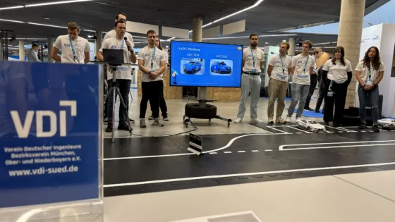 Präsentationsszene der HS München bei der VDI Autonomous Driving Challenge 2025, powered by MdynamiX. Im Vordergrund steht ein Acrylaufsteller mit dem VDI-Logo und der Aufschrift „Verein Deutscher Ingenieure Bezirksverein München, Ober- und Niederbayern e.V.“. Dahinter befindet sich eine markierte Teststrecke mit einem Schild „PIT OUT“ und einem Modellfahrzeug. Eine Gruppe von Teilnehmenden steht neben einem großen Bildschirm, der eine Folie mit dem Titel „AADC Platform“ zeigt, inklusive Bildern von zwei Modellfahrzeugen mit den Bezeichnungen „AADC 2024“ und „AADC 2025“.