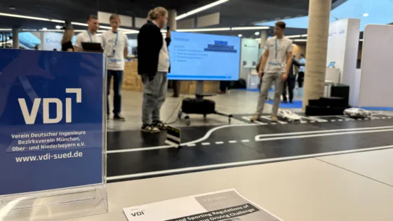 Szene bei der VDI Autonomous Driving Challenge 2025, powered by MdynamiX. Im Vordergrund steht ein Acrylaufsteller mit dem VDI-Logo und der Aufschrift „Verein Deutscher Ingenieure Bezirksverein München, Ober- und Niederbayern e.V.“. Daneben liegt ein Dokument mit dem Titel „Technical and Sporting Regulations of the Autonomous Driving Challenge“. Im Hintergrund befindet sich eine Präsentationsfläche mit markierten Fahrspuren, kleinen Modellfahrzeugen und einem großen Bildschirm, auf dem technische Inhalte angezeigt werden. 