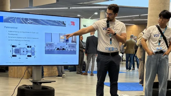 Präsentation bei der VDI Autonomous Driving Challenge 2025, powered by MdynamiX. Eine Person der Teams der TU Berlin steht vor einem großen Bildschirm und erklärt Inhalte zu Parkmanövern. Auf dem Bildschirm sind technische Grafiken und Text sichtbar, darunter die Überschrift „Parkmanöver“ sowie Punkte wie „Implementierung von Algorithmen für Quer- und Parallelparken“ und „Dynamische Anpassung der Zeitfenster basierend auf der Entfernung zum Parkplatz“. 