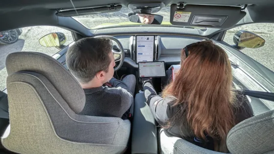 Testfahrer bewertet ein gefahrenes Fahrmanöver im ADAS-Benchmark mit der MXevalApp auf einem Tablet, das ihm von einer Mitinsassin überreicht wird.